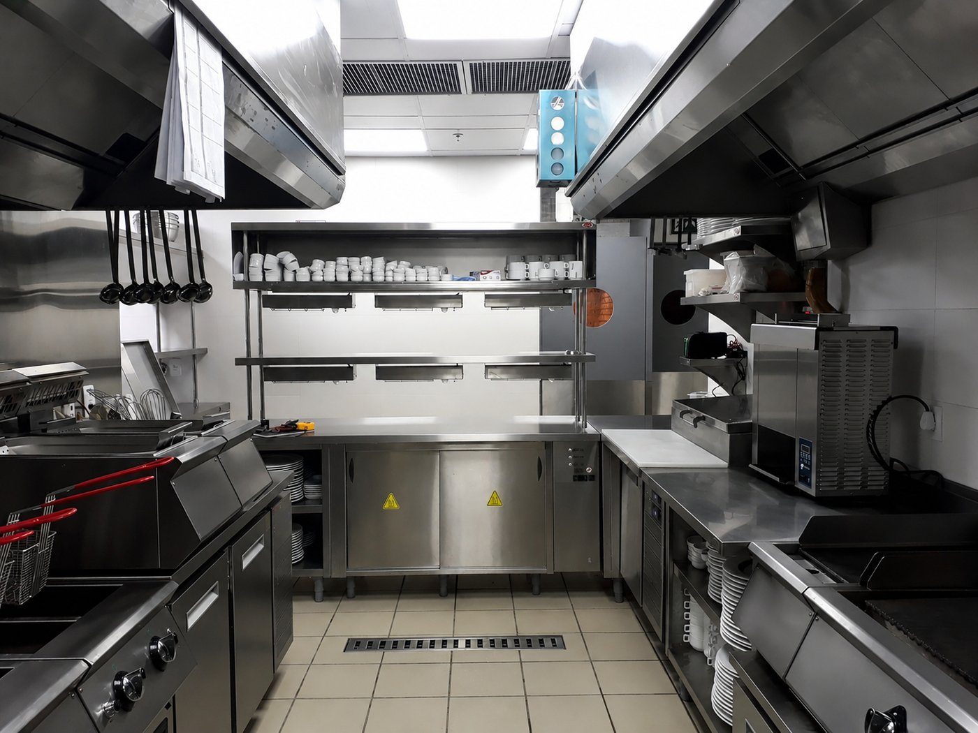 Cozinha profissional em operação com fritadeiras, bancadas em inox, prateleiras suspensas e coifas